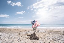 Rear view of young woman walking toward sea with USA flag on sandy beach. — Stock Photo
