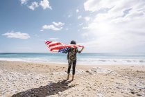 Rückansicht einer jungen Frau, die mit US-Flagge am Sandstrand steht. — Stockfoto