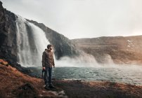 Вид сбоку на человека, стоящего с фотокамерой и отводящего взгляд на большой водопад в Исландии. — стоковое фото