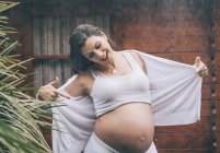 Smiling pregnant woman pointing on belly in rain against wooden house — Stock Photo