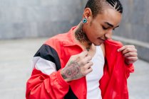 Smiling hipster with piercing and tunnel in ears holding red casual jacket in hands on grey blur background — Stock Photo