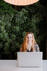 Smiling pretty redhead woman using laptop at table against bush — Stock Photo