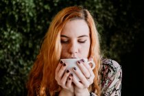 Rossa giovane donna che beve dalla tazza contro il cespuglio — Foto stock