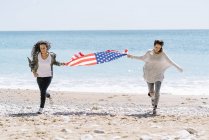 Zwei junge erwachsene Frauen laufen mit US-Flagge am Strand. — Stockfoto