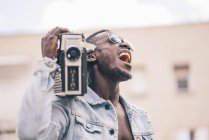 Smiling black man in sunglasses walking with vintage radio device and singing — Stock Photo