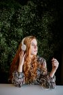 Jeune rousse femme avec écouteurs assis à table contre la brousse — Photo de stock