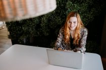 Cheerful young redhead woman using laptop at table — Stock Photo