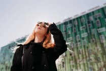 Стильна молода руда жінка в сонцезахисних окулярах дивиться на камеру на зеленому фоні . — стокове фото
