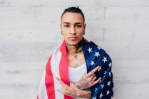 Латиноамериканське татуювання людини з пірсингом обгортаючи себе прапором об'єднаних станів і дивлячись на камеру на сірому текстурованому тлі — стокове фото