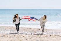 Две взрослые женщины бегут по пляжу с флагом США
. — стоковое фото