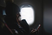 Hübsche Frau blickt auf das Handy, das im Flugzeugfenster sitzt. — Stockfoto