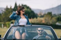 Brunette femme en jean décontracté vêtements assis dans la voiture avec barbu homme en lunettes de soleil — Photo de stock