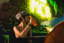 Стильна молода азіатка сидить у кафе і п'є пляшку — стокове фото