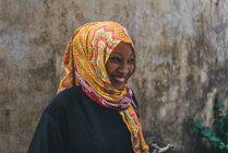 CAMERÚN - ÁFRICA - 5 DE ABRIL DE 2018: Joven mujer africana sonriente con el tocado brillante de pie en la pared áspera . - foto de stock