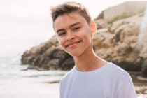 Portrait de mignon garçon souriant en t-shirt blanc debout sur le bord de la mer en été — Photo de stock