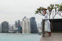 Pretty woman in bathrobe standing at pool in city with skyscrapers on background — Stock Photo