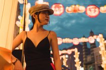 Jeune femme asiatique à la mode regardant loin dans la ville illuminée dans la soirée — Photo de stock