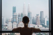 Mujer de pie en casa y mirando a fondo la ventana de los rascacielos - foto de stock
