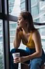 Thoughtful young woman with cup sitting in apartment and looking through window — Stock Photo