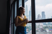 Усміхнена красива азіатська жінка стоїть з чашкою на вікно і дивиться геть — стокове фото
