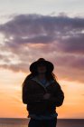 Woman in hat and jacket standing at evening sky on coast — Stock Photo