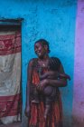 КАМЕРУН - Африка - 5 апреля 2018 года: Улыбающаяся взрослая африканская женщина, держащая обнаженного ребенка и стоящая у красочной стены на улице — стоковое фото