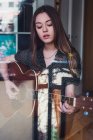 Jovem mulher bonita de pé na janela e tocando guitarra acústica em casa — Fotografia de Stock