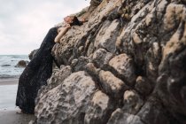 Mulher na moda em vestido preto encostado na rocha na praia — Fotografia de Stock