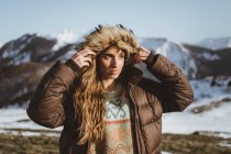 Attractive woman putting on hood of warm jacket and looking away in snowy mountains — Stock Photo