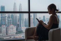 Азіатська жінка зі смартфоном сидить у кріслі в квартирі з видом на місто — стокове фото