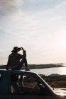 Жінка в капелюсі сидить на даху автомобіля на морі і дивиться на погляд — стокове фото