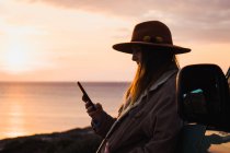 Femme en chapeau en utilisant un smartphone tout en s'appuyant sur la voiture au coucher du soleil — Photo de stock