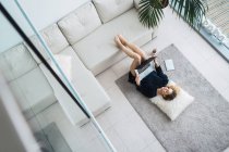 Jeune femme couchée sur le sol avec les jambes sur le canapé et le surf ordinateur portable — Photo de stock