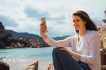 Женщина делает селфи со смартфоном на скалистом побережье — стоковое фото