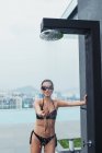 Pretty cheerful woman in swimwear standing at pool shower with city view on background — Stock Photo