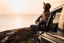 Frau genießt Sonnenuntergang und spielt Gitarre, während sie am Meer im Auto sitzt — Stockfoto