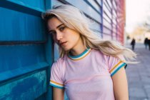 Junge blonde Frau blickt in die Kamera, während sie sich an eine blaue Wand lehnt — Stockfoto