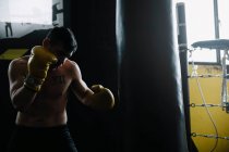 Boxer sem camisa em luvas de pé e saco de perfuração durante o exercício. — Fotografia de Stock