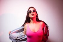 Jolie jeune femme rose habillée en lunettes de soleil debout au mur blanc et regardant la caméra — Photo de stock