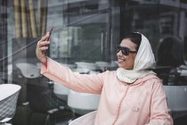 Mujer marroquí con hijab y vestido árabe típico tomando selfie en la cafetería - foto de stock