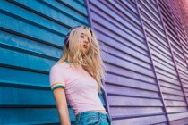 Молода блондинка стоїть на яскравій різнокольоровій стіні і слухає музику з фіолетовими навушниками — стокове фото