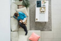 Frau im blauen Pullover sitzt auf Couch im stilvollen Wohnzimmer und liest Buch — Stockfoto
