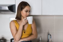 Вдумлива молода жінка з чашкою, що стоїть на кухні — стокове фото