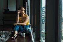 Женщина с чашкой сидит на полу дома и смотрит в окно — стоковое фото