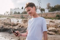 Boy with smartphone smiling while standing on rocky coast — Stock Photo
