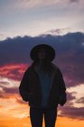 Вдумчивая женщина в шляпе и куртке, стоящая на закате под драматическим небом — стоковое фото