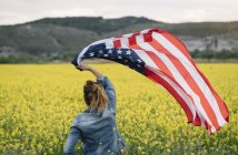 Mulher segurando bandeira americana no campo com flores amarelas no Dia da Independência — Fotografia de Stock