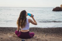 Fit woman sitting on shore at ocean and drinking water from bottle — Stock Photo