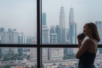 Lächelnde Frau spricht mit Smartphone am Fenster in Wohnung mit Blick auf die Stadt — Stockfoto