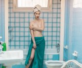 Junge Oben-ohne-Frau in Handtücher gehüllt, blickt in blaues Badezimmer in die Kamera — Stockfoto
